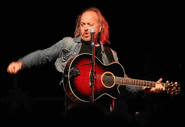 Bill Bailey performing on stage holding a guitar and starting in front of a microphone.  