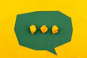 Yellow sheets of lined paper lie on a green chalk board and form a chat bubble with three crumpled papers in the center..