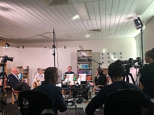 A room full of people filming a video. Four people are being recored like a round table discussion. Two other people sit off to the side watching a monitor.   